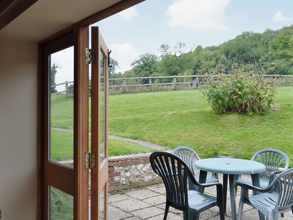 Une porte ouverte donne sur un patio équipé d’une table et de chaises, un gazon verdoyant et une vue sur une colline parsemée d’arbres.