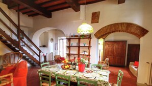 Un comedor con una mesa redonda dispuesta para seis personas, rodeada de sillas verdes. Un candelabro cuelga encima, y se ve una escalera de madera a la izquierda.
