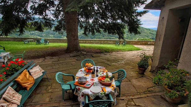 Un área de comedor al aire libre con una mesa de piedra preparada para una comida, rodeada de sillas verdes y un banco con cojines.