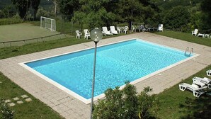 Una piscina rectangular con superficie de agua azul cristalina, rodeada por un solárium empedrado. Varias sillas de descanso blancas están dispuestas alrededor de la piscina, y se ve un arco de fútbol al fondo.
