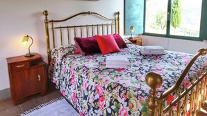 Een slaapkamer met een bloemenbedekking, een messing bedframe, een nachtkastje met lamp, en een raam met uitzicht op groen.