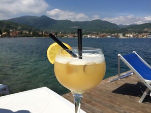 A glass of drink with a lemon slice and straw, placed on a table with a view of a lake and mountains in the background. - Holiday Home in Manerba del Garda Near Lake (Manerba del Garda)