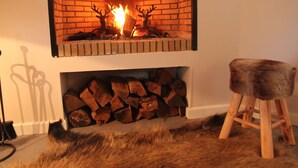Ein Kamin mit Ziegelverkleidung und Kaminabdeckung, ein Holzstoß darunter und ein hölzerner Hocker mit Fellauflage rechts.