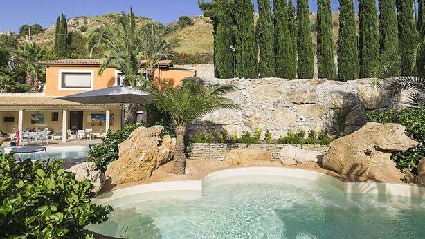 Zona de piscina con muro de piedra, palmeras y un edificio con asientos al aire libre.