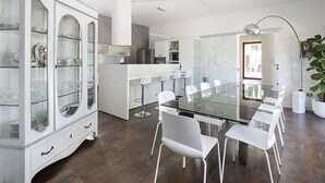 Una cocina moderna con una mesa de comedor de vidrio y sillas blancas, un armario blanco con puertas de vidrio, y una planta grande.