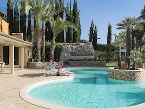 A swimming pool with a stone border, surrounded by palm trees and a stone wall. - Exclusive Luxury Villa in Agrigento with Private Pool, Hot Tub, BBQ (Agrigento)