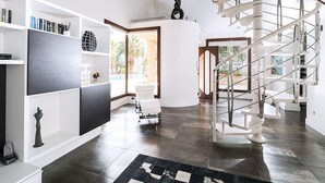 Un interior moderno con una escalera espiral, un ventanal con vistas a palmeras y una estantería llena de libros y objetos decorativos.