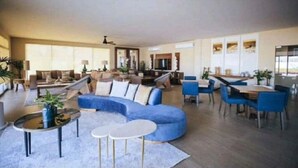 A spacious living area with a large blue sectional sofa, a dining table with blue chairs, and a kitchen area in the background. - OCEAN VIEW AMAZING CONDO@VISTAVELA4104 (Cabo San Lucas)