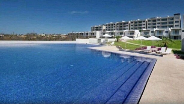 A large swimming pool with a clear blue water surface, surrounded by a concrete deck. In the background, there is a multi-story hotel building with balconies and a grassy area with umbrellas. - OCEAN VIEW AMAZING CONDO@VISTAVELA4104 (Cabo San Lucas)