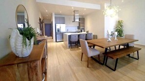 A modern kitchen with a central island, a dining area with a wooden table and benches, and a large mirror on the left. - OCEAN VIEW AMAZING CONDO@VISTAVELA4104 (Cabo San Lucas)