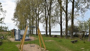 A playground with a swing set and a play structure, surrounded by trees and a body of water. - Ferienhaus mit Sauna in Boiensdorf am Salzhaff (Boiensdorf)