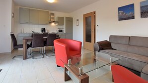 A modern living room with a glass coffee table, a red armchair, and a grey sofa. A kitchen with white cabinets and a microwave is visible in the background. - Ferienhaus mit Sauna in Boiensdorf am Salzhaff (Boiensdorf)
