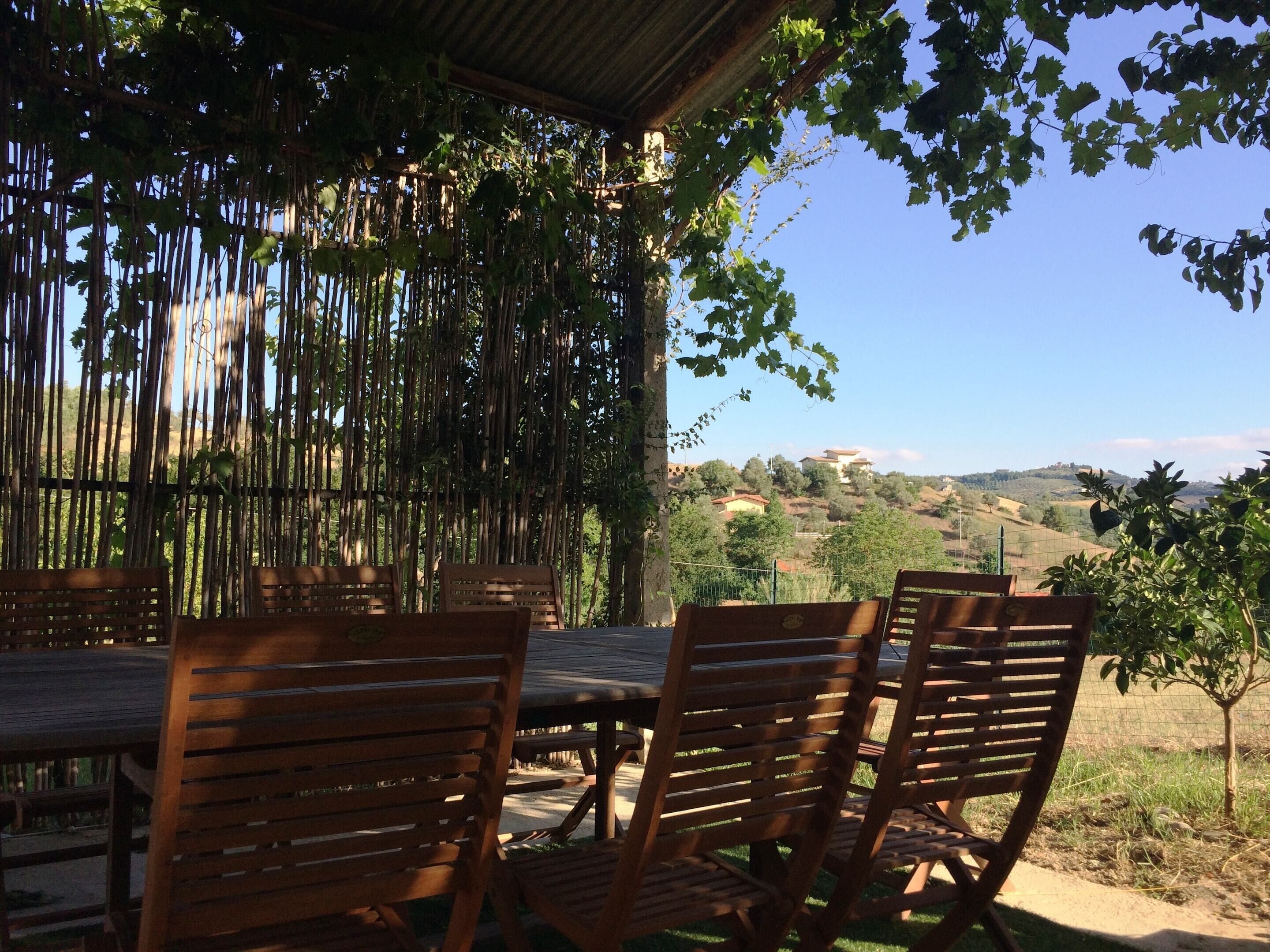 A wooden table and chairs under a bamboo structure with a view of a hilly landscape.