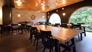 Un vaste espace dédié à la salle à manger, avec une longue table en bois entourée de chaises, un mur de briques orné d’une horloge décorative et une grande fenêtre en arc offrant une vue sur la verdure.