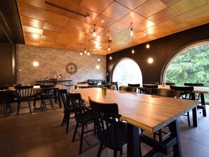 A spacious dining area with wooden tables and chairs, a brick wall with a clock, and a large arched window offering a view of greenery. - Spacious Holiday Home in Vielsalm near Baraque de Fraiture (Vielsalm)