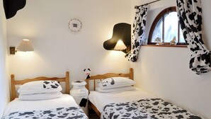 A room with two beds, each with a cow-print bedspread, a bedside lamp, a wall clock, and a window with cow-print curtains. - The Coach House - 15661 (East Tytherton, Chippenham)