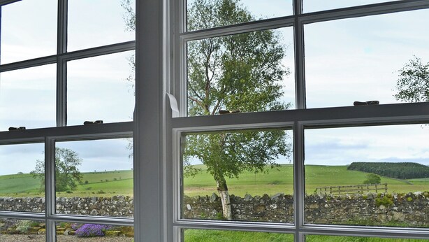 A view through a window with multiple panes, revealing a rural landscape with green fields, a stone wall, and a tree. - 2 bedroom accommodation in Alnham, near Rothbury (Alnham, near Rothbury)