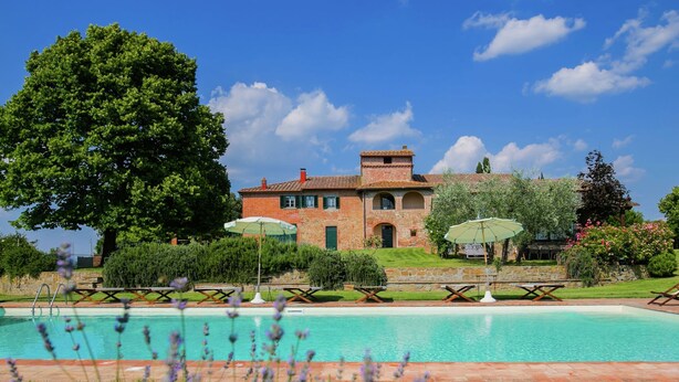 Zona de piscina con sillas de descanso y sombrillas, un árbol grande y un edificio de piedra con tejado de baldosas.