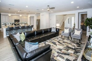 A spacious living room with a black leather sectional sofa, grey armchairs, and a patterned rug. A kitchen with white cabinets and a dining area with bar stools is visible in the background. - 2606YA Luxurious En-Suite Single Home Pool (Kissimmee)