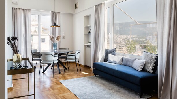 Una sala de estar moderna con un sofá azul, una mesa de comedor, sillas y vistas de un paisaje urbano a través de grandes ventanales.