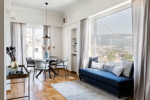 A modern living room with a blue sofa, dining table, chairs, and a view of a cityscape through large windows.