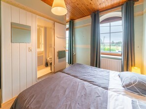 A bedroom with a large bed, a window with curtains, a wooden ceiling, and a bathroom with a toilet and sink. - Former Hotel with Swimming Pool, Hot Tub, Sauna for Family Groups (Malmedy)