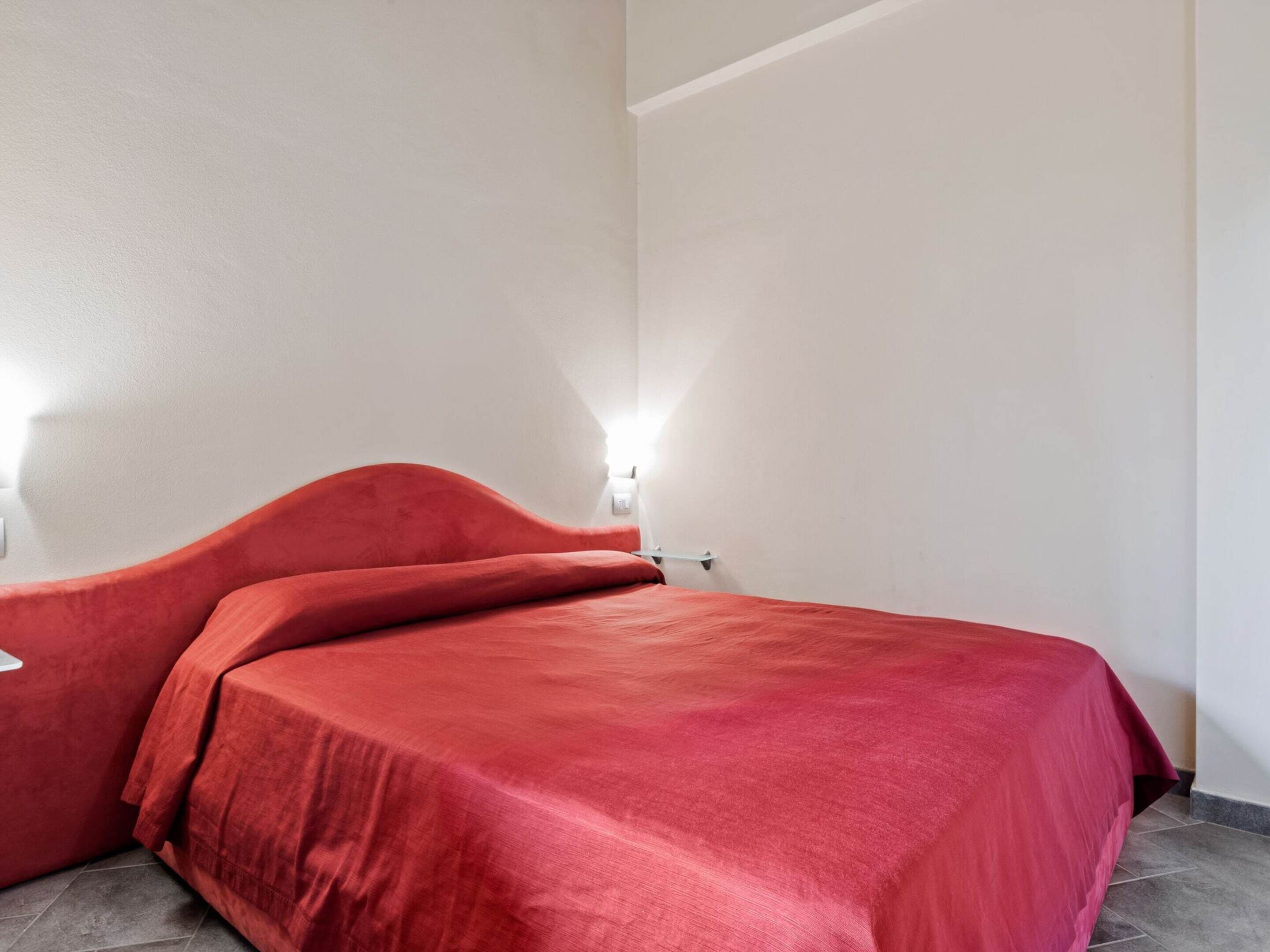 A room with a bed covered in a red blanket, a white wall, and a light switch.
