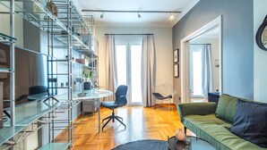 A modern living room with a green sofa, a round coffee table, and a glass dining table with chairs. There is a metal shelving unit with various items on it, and a large window with curtains. - Superb 1BD Apartment in the heart of Kolonaki (Athens)