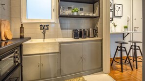 A modern kitchen with a built-in oven, a sink, and a window above the countertop. There are shelves with glassware and a small plant. - Superb 1BD Apartment in the heart of Kolonaki (Athens)