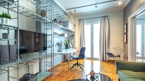 A modern living room with a sofa, a coffee table, and a TV. There are shelves with various items and a chair. - Superb 1BD Apartment in the heart of Kolonaki (Athens)
