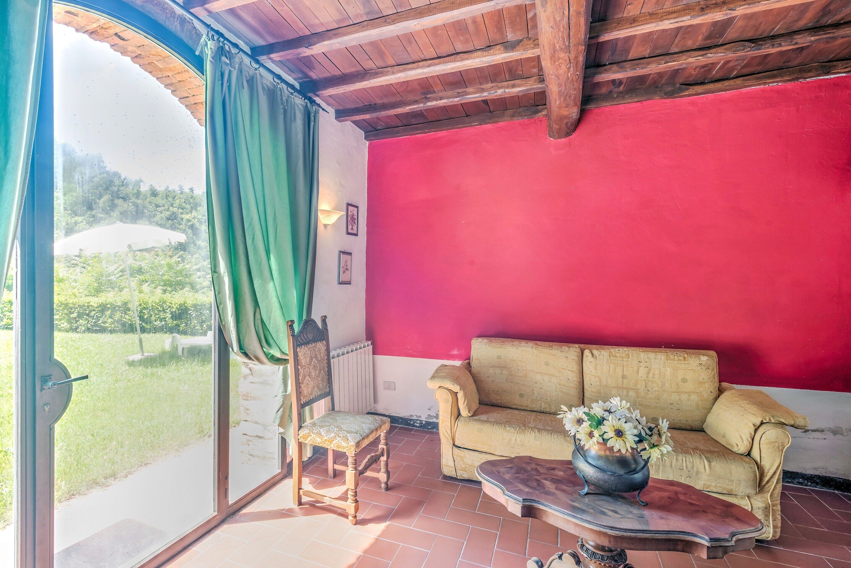 A room with a red wall, a beige sofa, a wooden chair, a small table with a vase, and a large window with a green curtain.