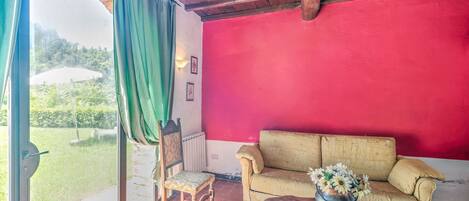 A room with a red wall, a beige sofa, a wooden chair, a small table with a vase, and a large window with a green curtain.