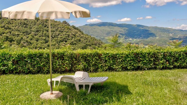 Un espace herbeux avec une chaise en plastique blanche et un parasol, entouré d’une haie et offrant une vue sur des collines ondulantes.