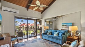 Ein geräumiges Wohnzimmer mit einem blauen Sofa, einem Holztisch und einem großen Fenster mit Blick auf eine tropische Landschaft.