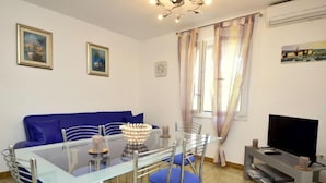 A modern living room with a glass dining table, blue chairs, a blue sofa, a flat-screen TV, and a decorative vase.