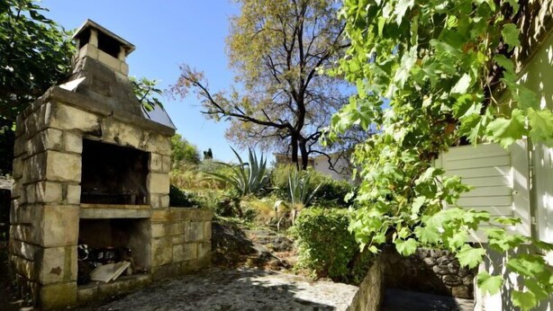 A stone barbecue grill in a garden with a stone wall and a tree.