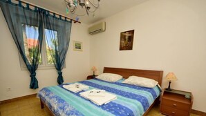 A bedroom with a bed, two bedside tables, a window with curtains, an air conditioning unit, and a painting on the wall.