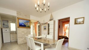 A dining area with a glass table and white chairs, a kitchen with a refrigerator, and a painting on the wall. - Villa Bay View by Interhome (Brač/Postira)