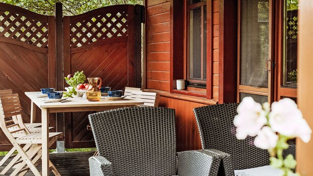 A wooden deck with a table set for dining, wicker chairs, and a window with a view of greenery.