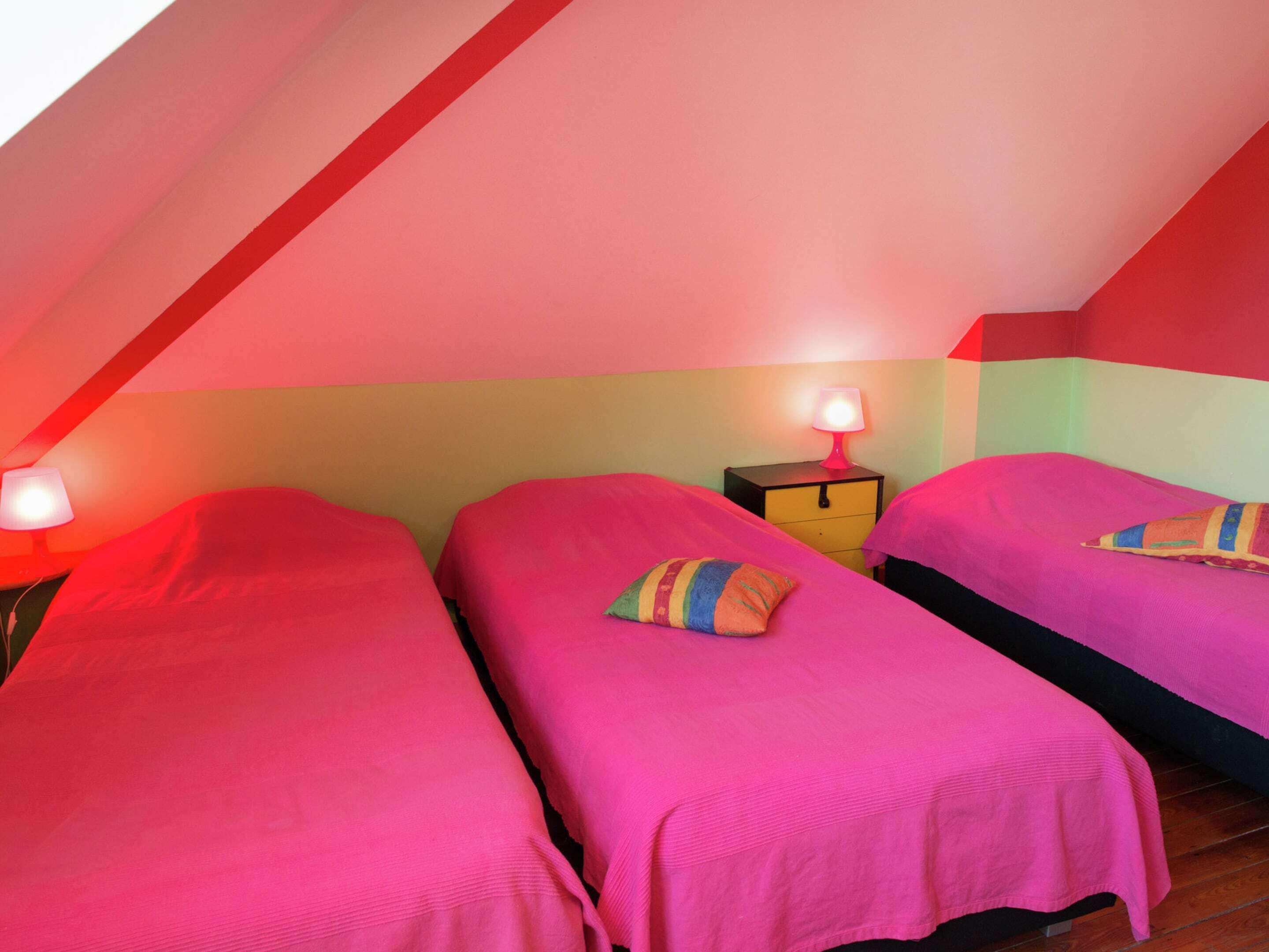 A room with two beds, each with a pink bedspread and a pillow. There is a nightstand with a lamp between the beds. The room has a sloped ceiling and a green wall.