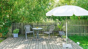 Eine Holzterrasse mit zwei Korbstühlen und einem kleinen Tisch, umgeben von üppiger Vegetation und einem Holzzaun.