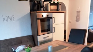 Eine moderne Küche mit Einbauofen und Mikrowelle, ein Holztisch mit Pflanze und Glas sowie eine Couch im Hintergrund.
