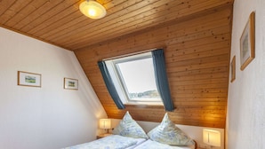 A bedroom with a wooden ceiling, a window with curtains, a bed with floral bedding, two bedside tables with lamps, and framed pictures on the wall. - Gemutliches Appartement in Hallenberg mit Terrasse (Hallenberg)