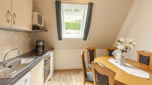 A compact kitchen with a microwave, sink, Bosch dishwasher, and wooden cabinets. There's a dining table with chairs and a vase of flowers. A window with curtains is visible, and the room has a sloped ceiling. - Gemutliches Appartement in Hallenberg mit Terrasse (Hallenberg)