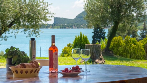 A table with a bottle, a bowl of bread, a plate of grapes, and two wine glasses, set against a scenic lake view. - Apartment in Manerba Near Lake Garda (Manerba del Garda)