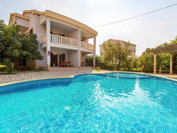 Ein Swimmingpool mit glatter, blauer Wasseroberfläche, umgeben von einem Gebäude mit Balkon und einer überdachten Terrasse.
