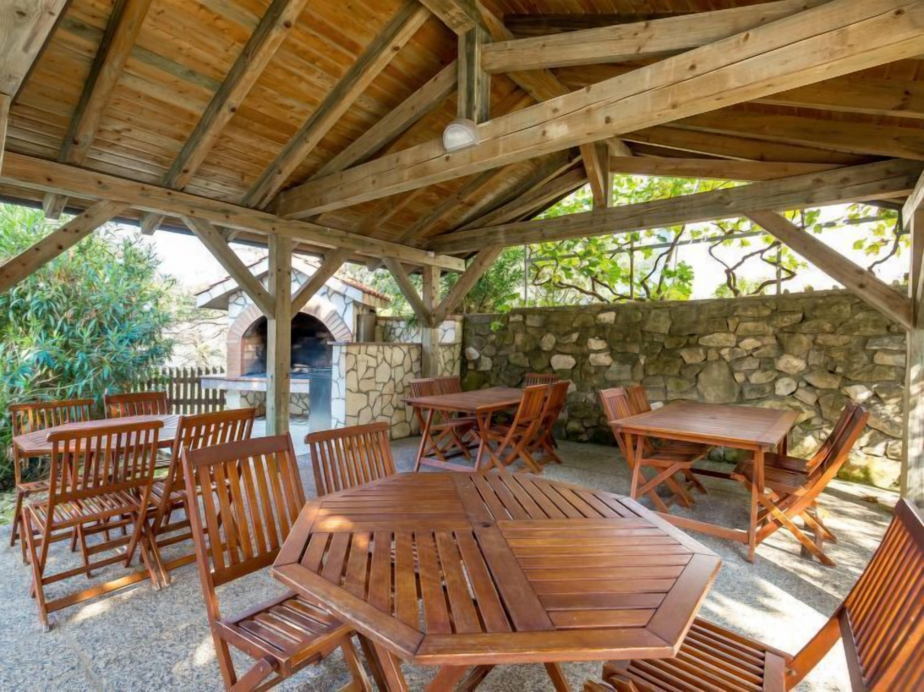 An outdoor wooden pavilion with stone walls and a stone oven, surrounded by wooden tables and chairs.