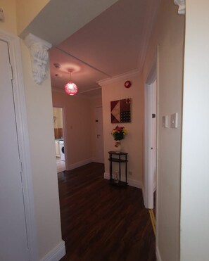 Un couloir doté d’un plancher en bois foncé, une porte blanche sur la gauche, un luminaire suspendu rouge et une petite table sur laquelle est posé un vase de fleurs.