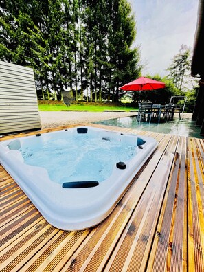 A hot tub on a wooden deck with a table and chairs under a red umbrella, surrounded by trees.