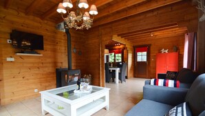 A cozy log cabin interior with a fireplace, a television, a dining area, and a living room with a sofa and cushions. - Chalet in Ardennes With Sauna & Garden (Waimes)